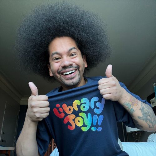 Depicts librarian Mychal Threets smiling widely with two thumbs up wearing a black shirt that says 'Library Joy!!' in rainbow letters.