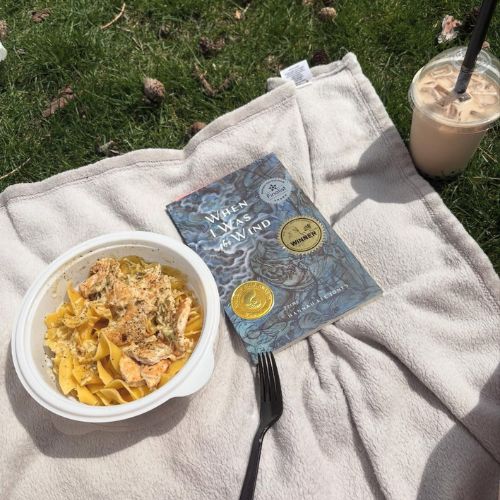 On a picnic blanket in the grass sits a bowl of food, a black plastic fork, and a copy of When I Was the Wind by Hannah Lee Jones, whose cover features a blue watercolor painting of birds. Next to the blanket is an iced drink.