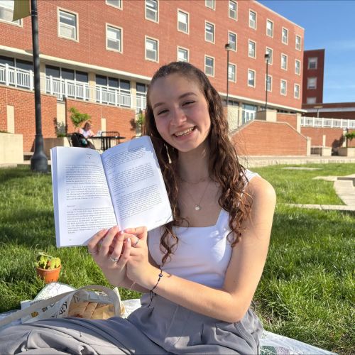 Solenn smiles as she poses in front of a red brick building, holding open a book with both hands with the pages facing toward the viewer. She sits on a blanket in the grass.