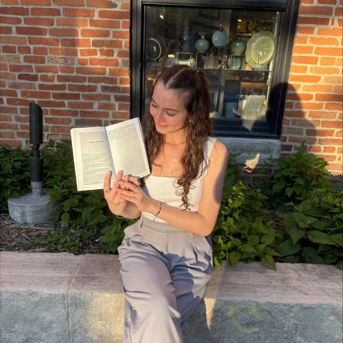 Solenn sits on the concrete ledge on the outside of a red brick building, in front of bushes, looking at the open pages of a book she is holding in both hands. A window with a black frame sits behind her.