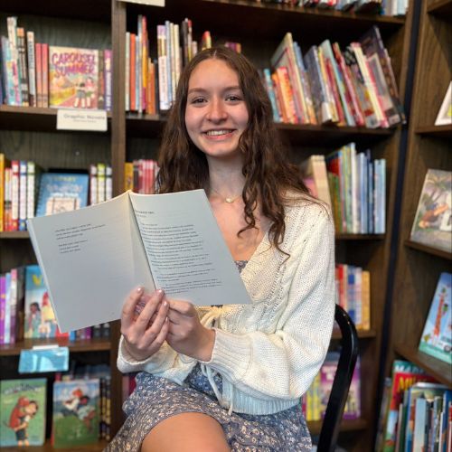 Solenn, an up-and-coming Bookstagrammer, smiles as she sits on a chair in front of full brown bookshelves, holding open a book that faces outward.