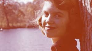 The poet Sylvia Plath as a young woman with her head against a tree, smiling in front of a body of water.