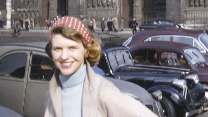 The poet Sylvia Plath with a polka dot head scarf and turtleneck sweater smiling in front of a row of parked cars.