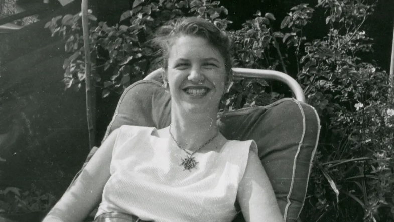 The poet Sylvia Plath as a young woman smiling widely sitting in an outdoor patio with her hair tied back in black and white.
