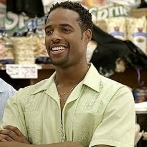 Movie still of actor Shawn Wayans smiling, wearing a yellow shirt, and with his arms folded.