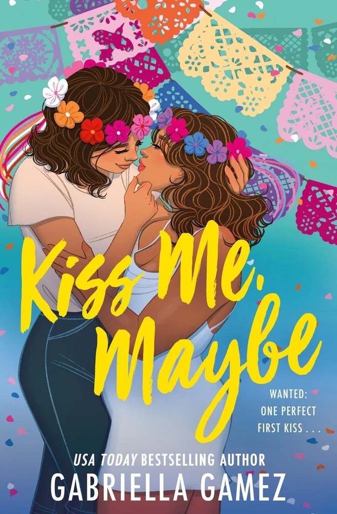 Two women lovingly embrace under colorful papel picado banners, wearing flower crowns. The bright cover reads "Kiss Me, Maybe" by Gabriella Gamez.