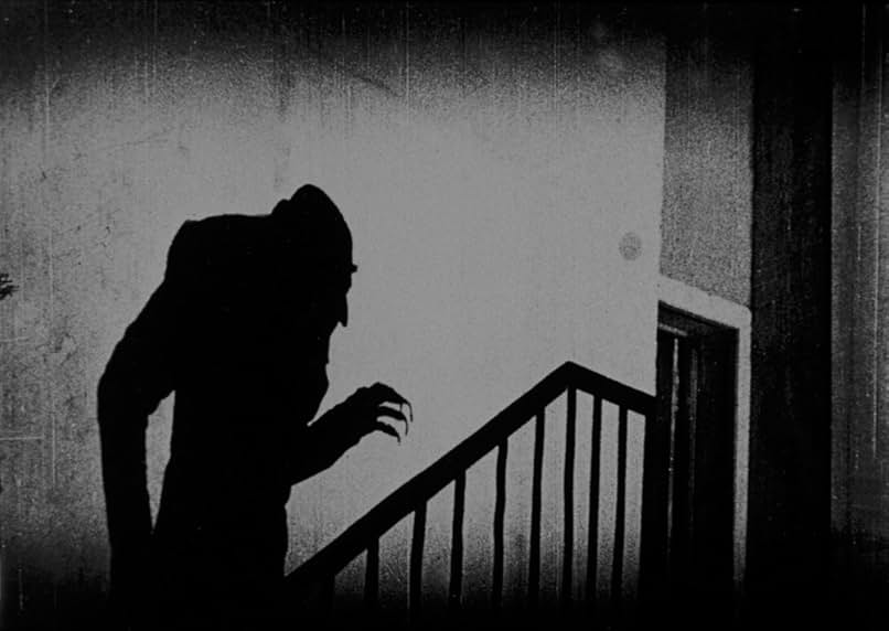 A still from the 1922 film Nosferatu. In black and white, the shadow of a hunched figure with long clawed fingers climbs a staircase.