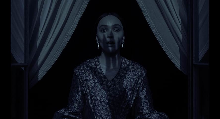A still from the 2024 film Nosferatu. A young woman dark floral Victorian dress stands at an open window in shadow with tears in her eyes. The shadow of a long clawed hand is over her face.