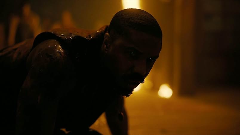 A still from the 2025 movie Sinners. A Black man with glowing eyes is on his hands and knees covered in blood. The background behind him is golden from a glowing fire.