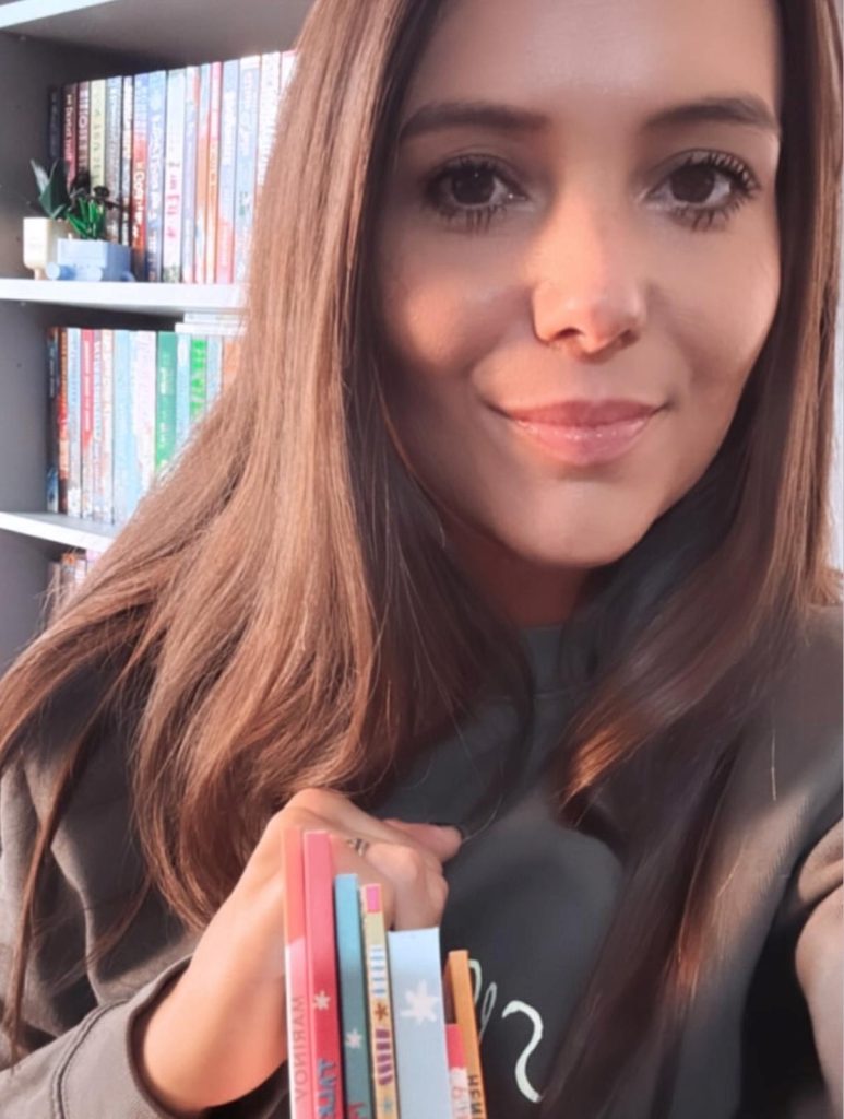 Bookstagrammer Hannah in a close up photo with a closed mouth smile. Peeking up from below are the bindings of multiple children's books in her hand as she stands in front of her colorful bookcase.