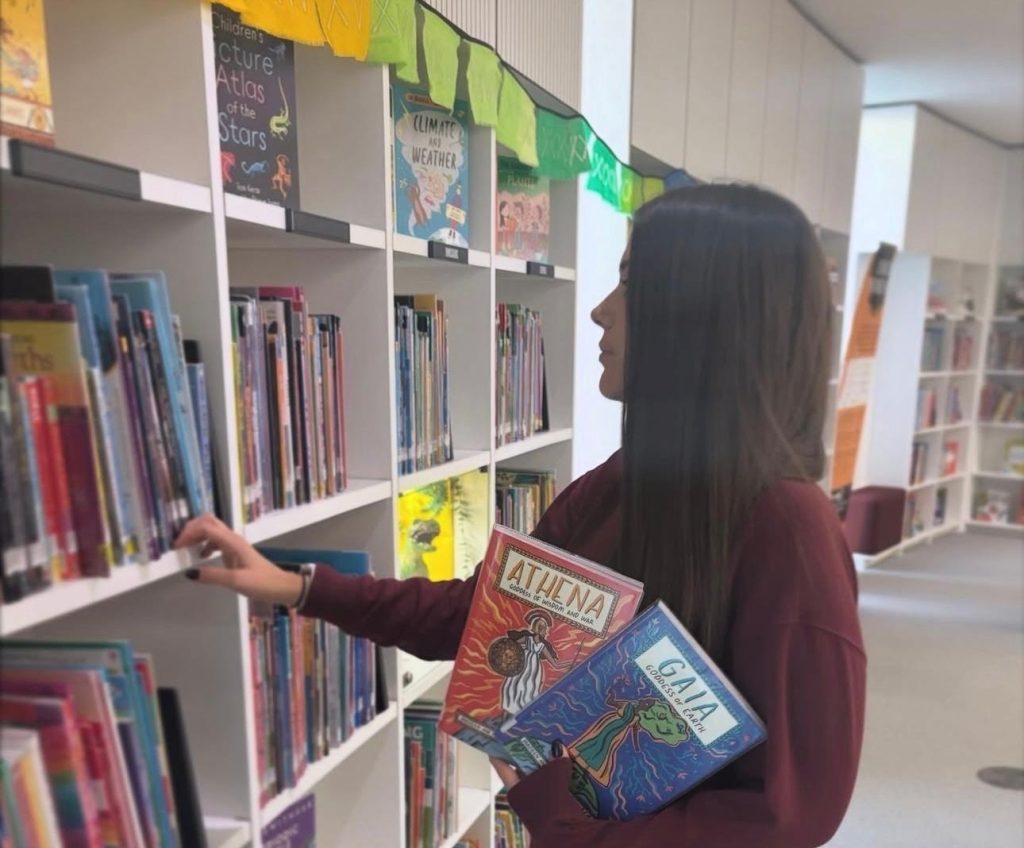 Bookstagrammer Hannah browsing the shelves of the library with two children's books, Athena and Gaia, in one arm while her other arm searches for other titles.