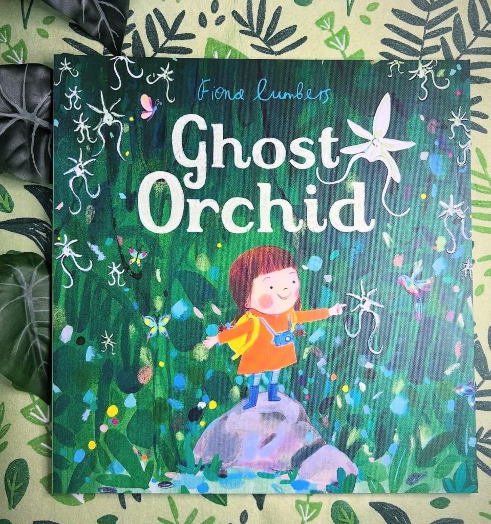 On a sage green table cloth decorated with different leaves, Fiona Lumber's children's book Ghost Orchid is displayed. The cover shows a small girl in a raincoat, rainboots, and a camera around her neck spreading her arms and smiling on top of a rock whilst surrounded by nature.