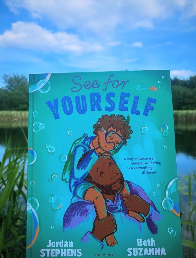 Bookstagrammer Hannah holds up See For Yourself by Jordan Stevens and Beth Suzanna in front of a pond on a sunny, blue-skied day. The blue book cover, spotted in air bubbles, features a boy in scuba gear on his father's shoulders as he gazes up at him.