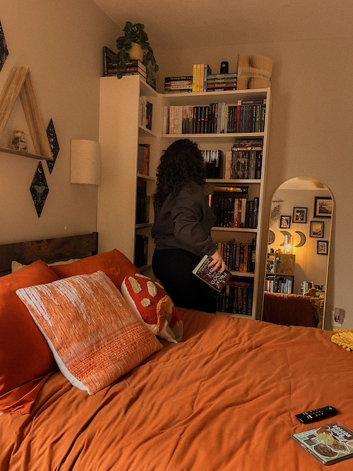 A girl facing away from the camera, holding a book in her hand. She is browsing the bookcase and an orange bedspread with a mushroom-shaped pillow is in front of her.