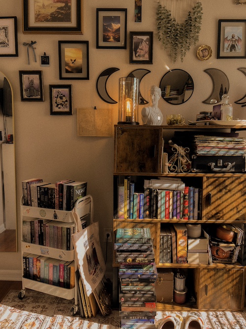 There is a rustic bookcase filled with numerous books. There is a statuette sitting atop the bookcase and multiple picture frames and mirrors on the wall above it. To the left, there is a small book cart filled with novels.