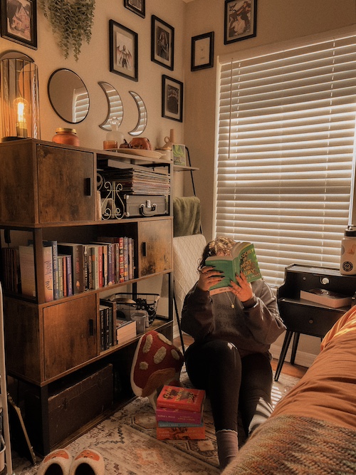 A girl is sitting on a carpet and reading A Court of Mist and Fury by Sarah J. Maas, a fantasy novel. Other novels from Sarah J. Maas' A Court of Thorns and Roses lay next to her.