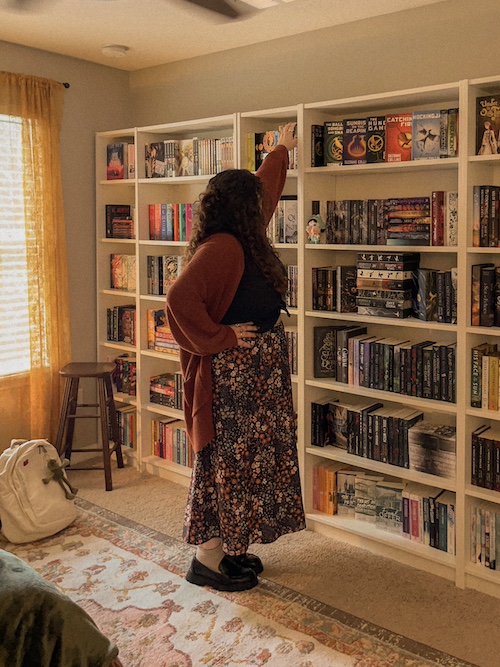 A girl reaching for a book at the top of her bookshelf. She is facing away from the camera.