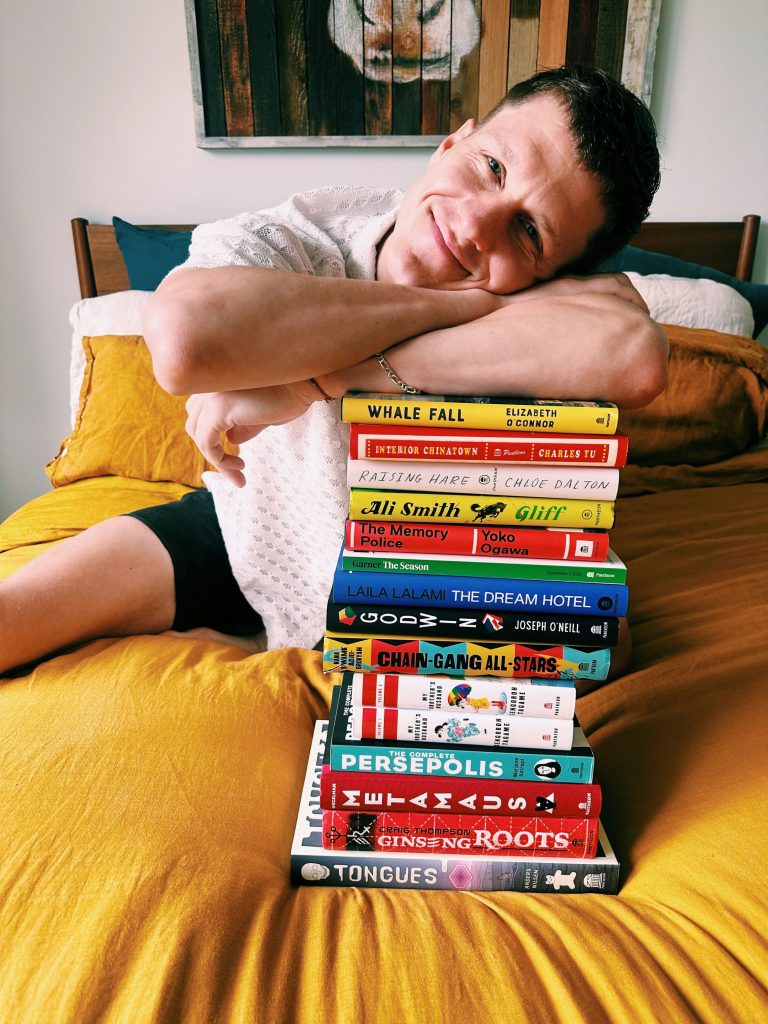 Bookstagrammer Christopher sits on a mustard colored duvet with his arms folded over a towering stack of books and his head tilted, resting on his bicep. Christopher smiles on top of his stack of 15 colorful books.