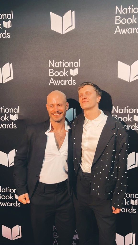 Bookstagrammer Christopher in a oversized black suit jacket with white pears, smiling while standing next to boyfriend and Bookstagrammer Bernie Lombardi at the National Book Awards.