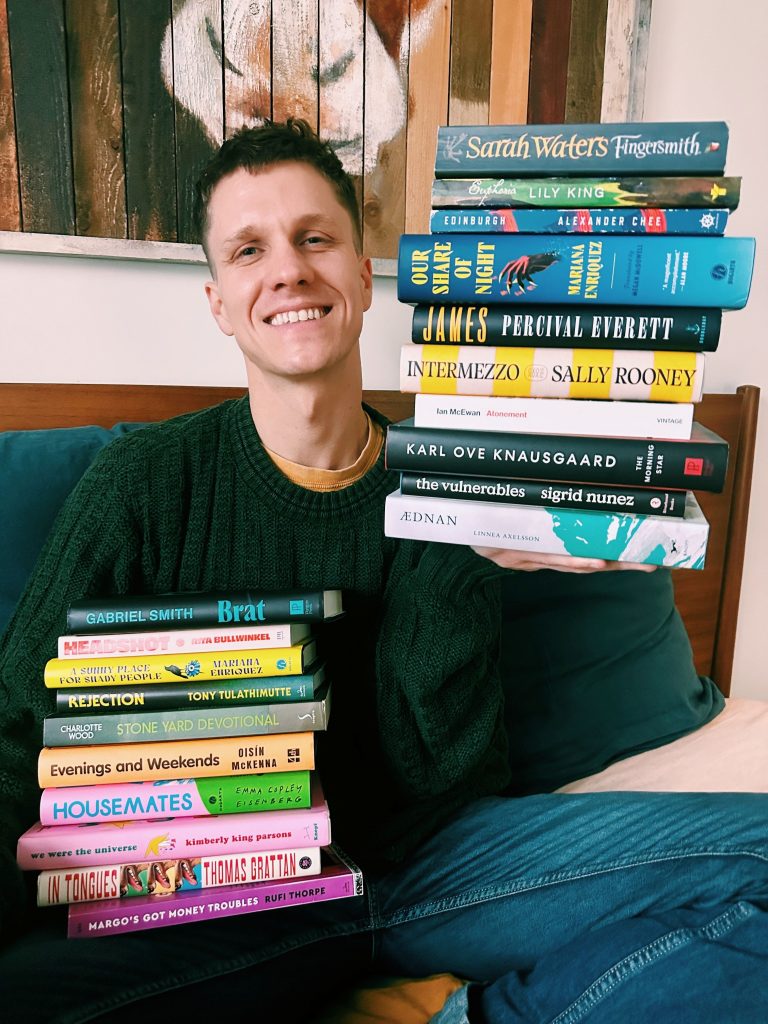 Christopher, sitting on his bed and wearing a forest green sweater, holds up one stack of books in his right hand (featuring novels by Sally Rooney, Marina Enriquez, Percival Everett, and more) and holds another stack in his lap (Featuring novels by Marina Enriquez, Charlotte Wood, Thomas Grattan, Oisin McKenna, and more).