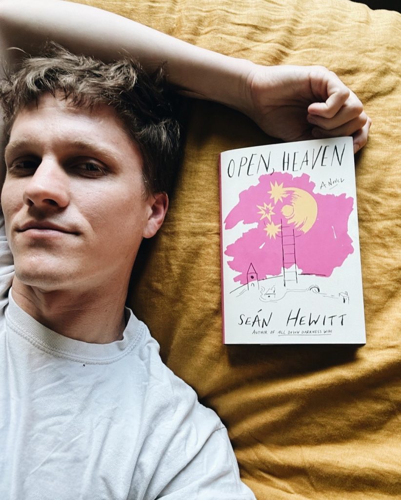Christopher, laying on mustard sheets, rests his head next to Seán Hewitt's novel Open, Heaven. His arm straddles over his head, his fingers lightly grazing the right corner of the novel.