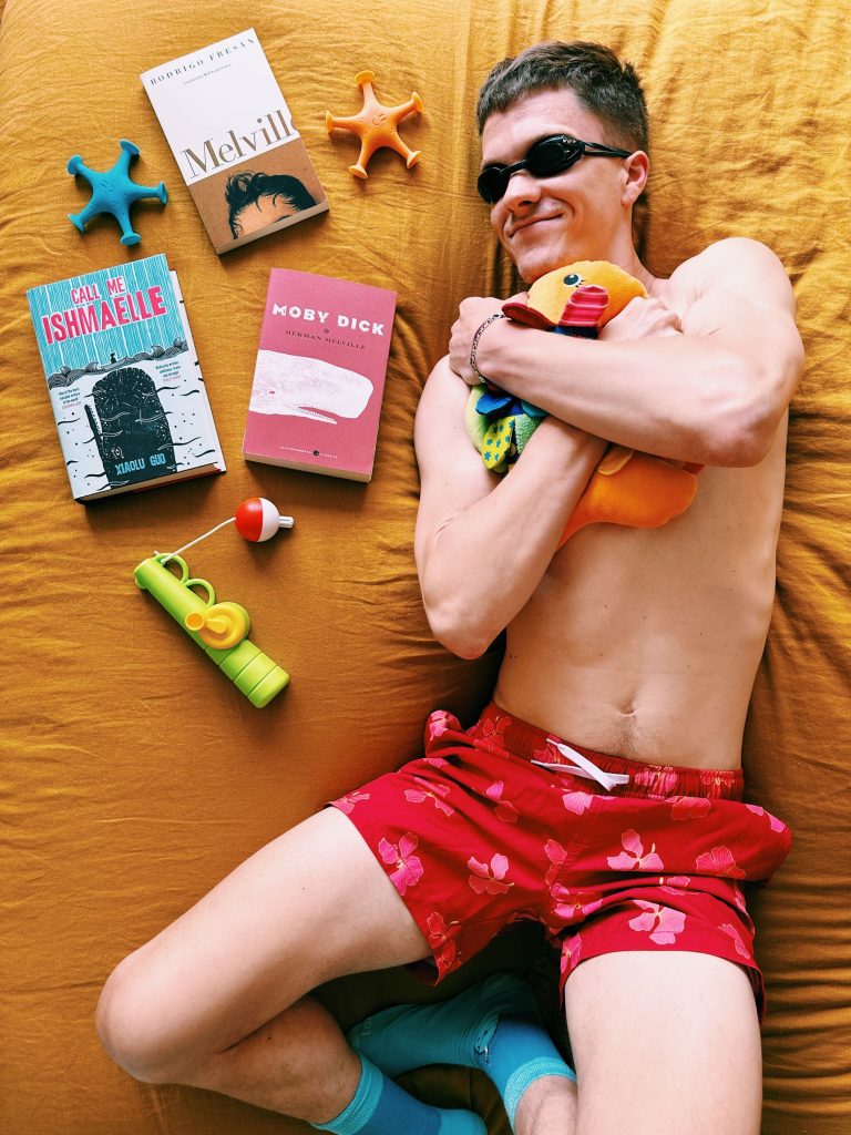 Christopher, laying on his mustard sheets, is in red swimsuit, clutching a fish stuffed animal, and wearing swimming goggles with a smile on his face. Next to him is a toy fishing rod and Moby Dick by Herman Melville, Call Me Ishmaelle by Xiaolu Guo, and a cut off Melville biography.