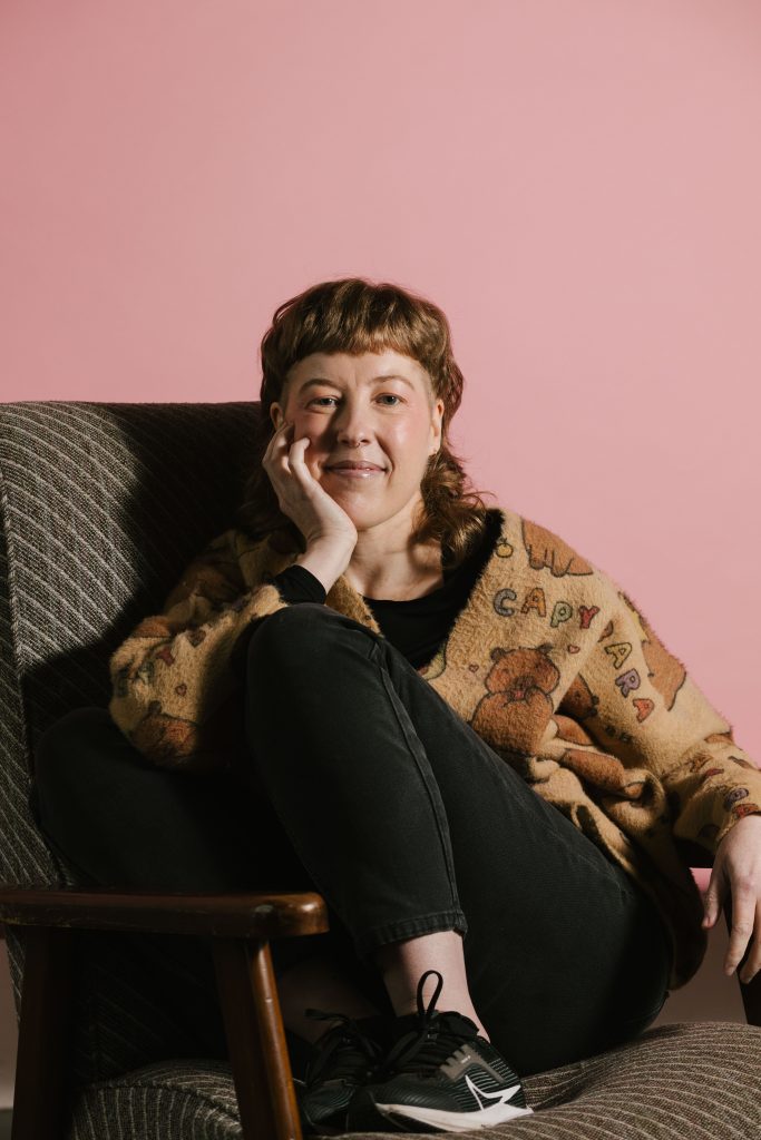 Trans author Elias Cold shitting on a chair, knees to check, with their hand holding their chin while wearing a capybara printed sweater and back jeans against a pink background.