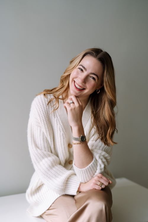 Joan O'Leary, a light-skinned woman with long dirty blonde hair, wearing a white sweater, sits and leans her chin on her hand, tilting her head to the viewer's right, smiling at the viewer. The background is gray.