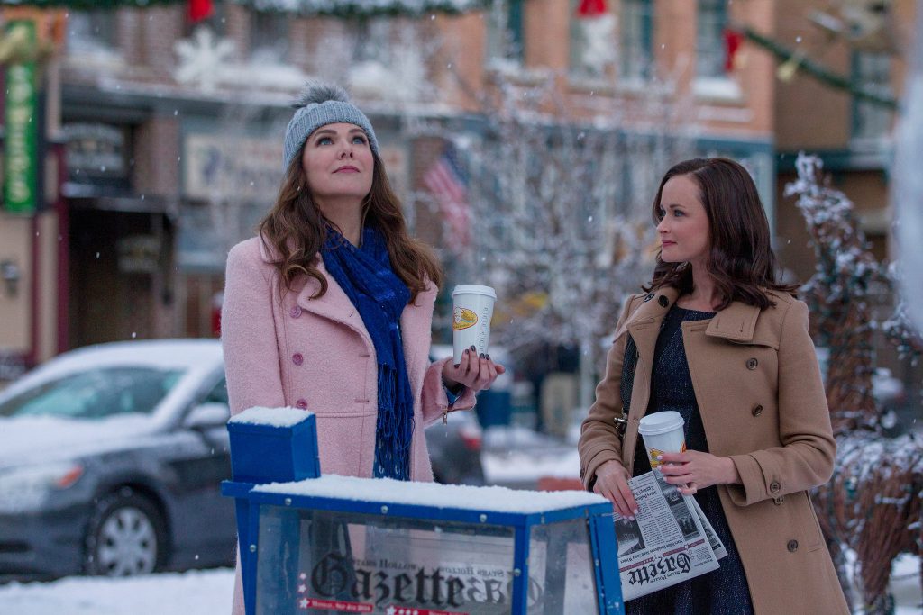 A still from the show Gilmore Girls: A Year in the Life. Two brunette woman bundled in winter coats stand outside on a small town street in the snow. Both women hold to-go coffee cups, and the one on the right also holds a newspaper.