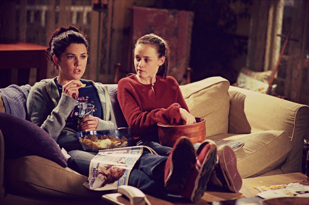 A still from the show Gilmore Girls. Two brunette women sit on a yellow couch and eat snacks out of bowls in their laps. A magazine rests on the legs of the woman on the left.