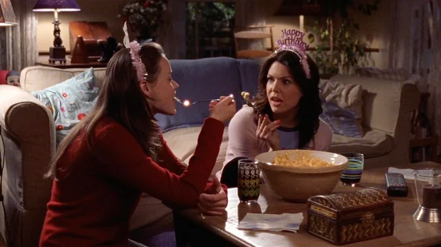 A still from the show Gilmore Girls. Two brunette woman wear pink "Happy Birthday" crowns and eat macaroni and cheese out of a large bowl set up on their coffee table. The two woman sit on the floor in front of their couch.