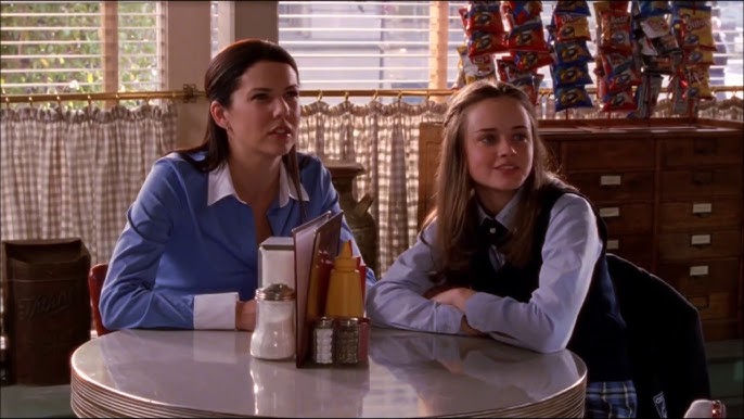 A still from the show Gilmore Girls. Two brunette woman, one wearing a blue private school uniform, sit at round table in a diner and stare off camera with bemused expressions.