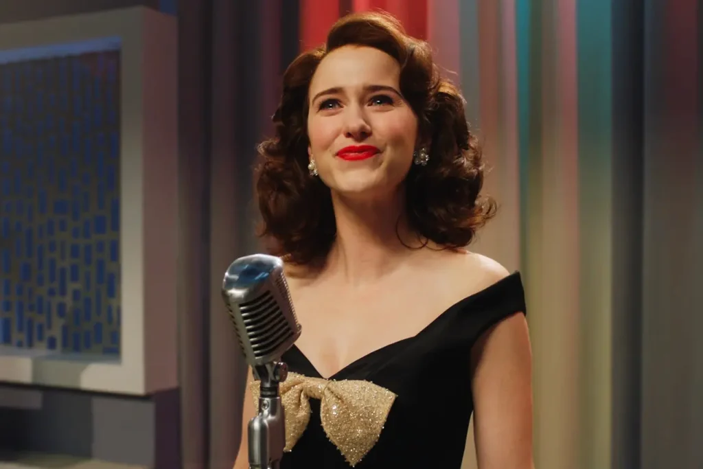 A still from the show The Marvelous Mrs. Maisel. A brunette woman with curly brown hair, a black dress, and costume jewelry, smiles in front of an old-fashioned silver microphone.