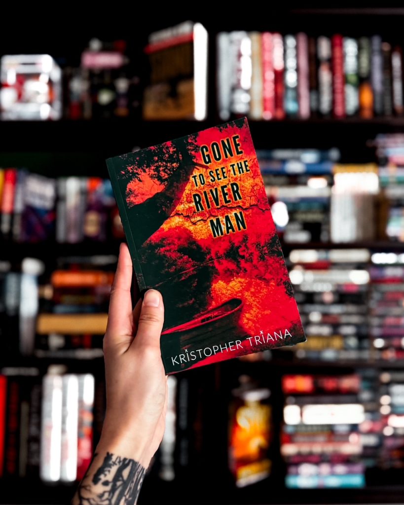 Depicts photo of Gone to See the River Man by Kristopher Triana book cover held up against a blurred background of a bookshelf.