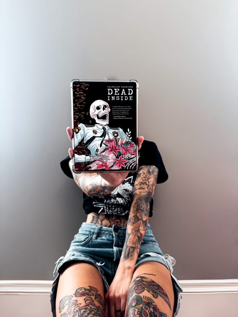 Depicts photo of Summer sitting and holding out an e-reader showing the cover of Dead Inside by Chandler Morrison. Book cover depicts a skeleton in a security guard uniform holding pink flowers