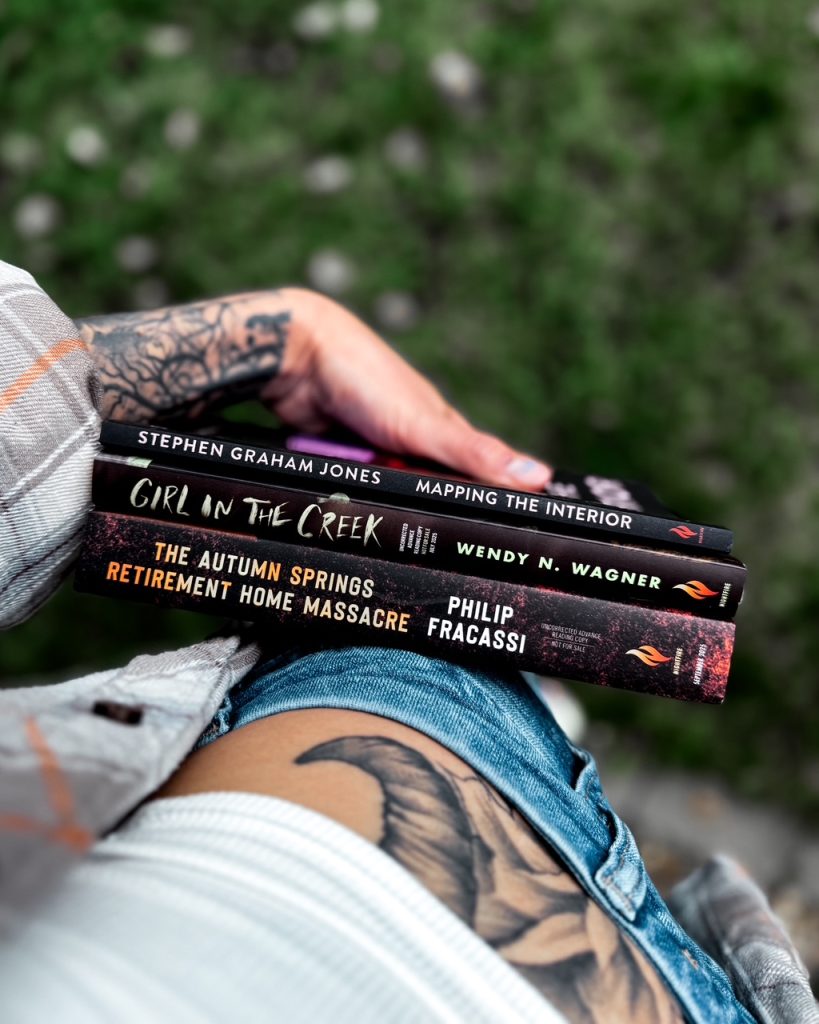 Depicts photo of the spines of Mapping the Interior by Stephen Graham Jones, Girl in the Creek by Wendy N. Wagner, and The Autumn Springs Retirement Home Massacre by Philip Fracassi. Person wears a gray flannel and jeans and is holding them towards a blurred ground.
