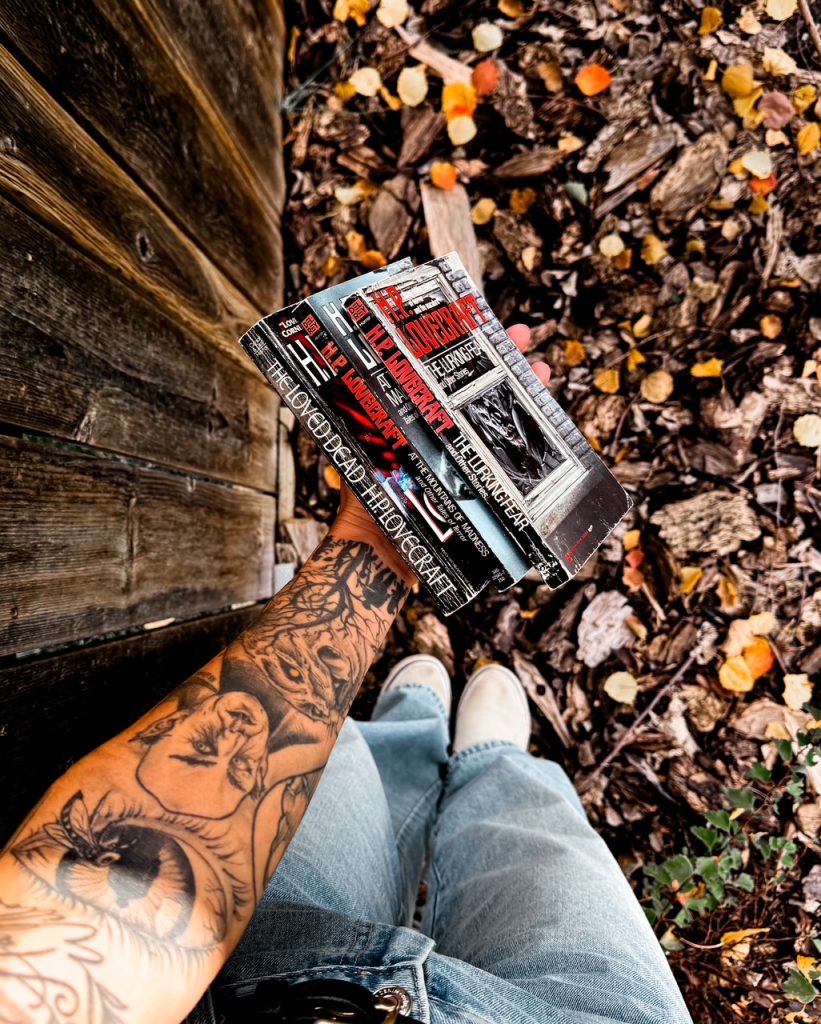 Depicts photo of the spines of three H.P. Lovecraft books held out against the ground with fall colored leaves and shows an arm with a sleeve of tattoos.