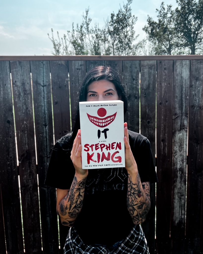 Depicts photo of Summer holding up the cover of It by Stephen King. The clown nose and red smile held up over her own face.