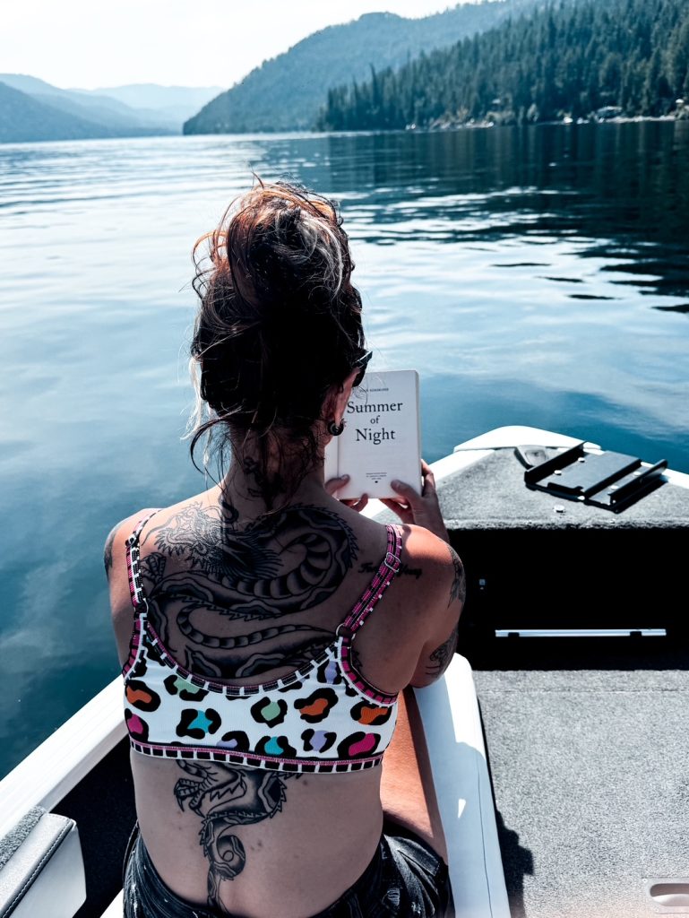 Image of the back of Summer with a cheetah print bathing suit and a messy bun reading Summer of Night in a boat out on the water.