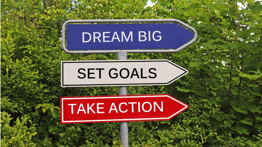 A three-directional sign in front of green foliage: "Dream Big" in blue, "Set Goals" in black, and "Take Action" in red, conveying motivation and determination.