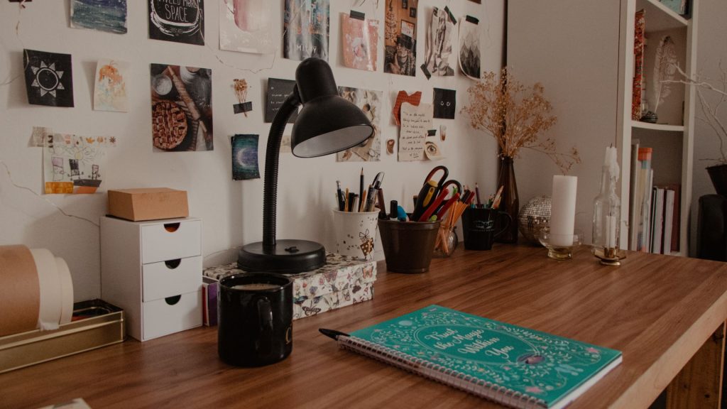 Cozy workspace with a wooden desk, black lamp, mug, notebooks, and art supplies. A wall collage of artwork and notes adds inspiration. Warm tones, creative vibe.