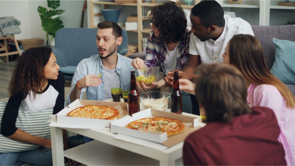 A group of six friends sit in a cozy living room, enjoying pizza and drinks. They are engaged in lively conversation, creating a warm, joyful atmosphere.