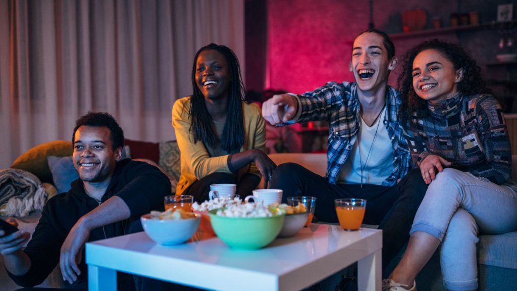 Four friends sit on a couch, laughing and pointing at a TV. A coffee table with drinks and popcorn is in front, a cozy atmosphere with warm lighting.