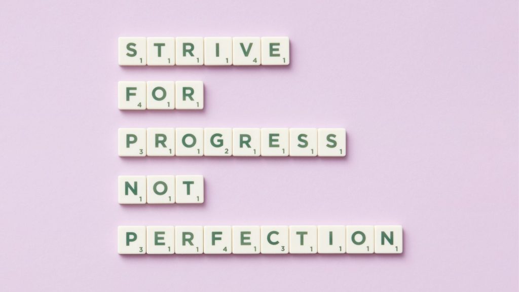 Text on Scrabble tiles spells "Strive for progress, not perfection" against a light purple background. The message inspires motivation and growth.