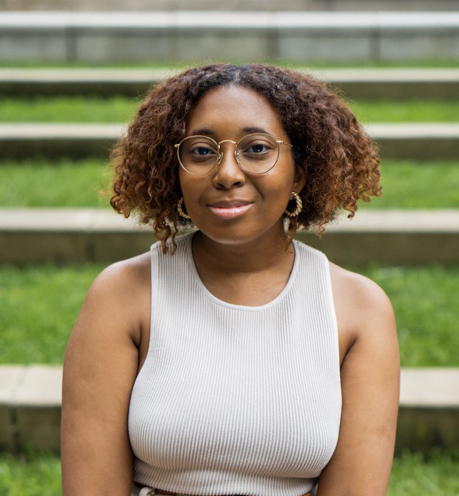 Author, Joelle Wellington, a Black woman, wearing a white tank top, skinny, golden framed, round glasses, and golden hoop earrings, smiling and looking ahead.
