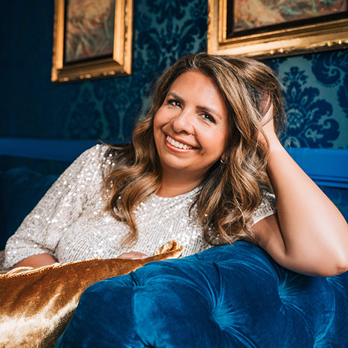 Headshot of author Nisha J Tuli. Tuli smiles and leans over the arm of a blue couch with her head resting on her hand.