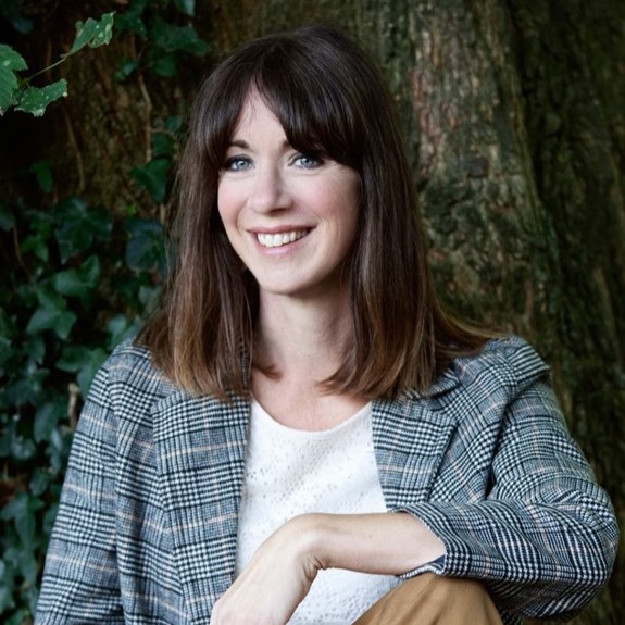 Cesca Major, a light-skinned woman with shoulder-length brown hair and a plaid jacket over a white shirt, smiles at the viewer while resting her arm on her knee. She sits in front of a tree.