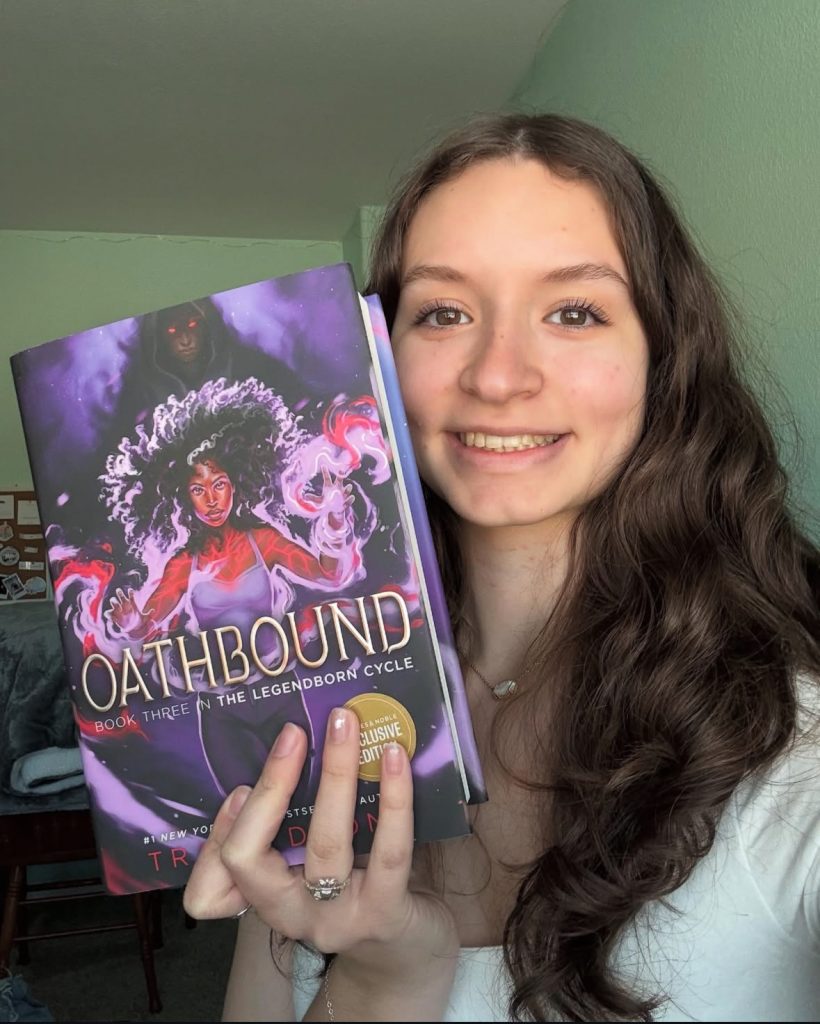 Solenn smiles as she poses for a selfie with a copy of Oathbound by Tracey Deonn, the cover of which features a Black woman with loose curly hair with swirls of magic around her hands and body with a hooded figure lurking behind her.
