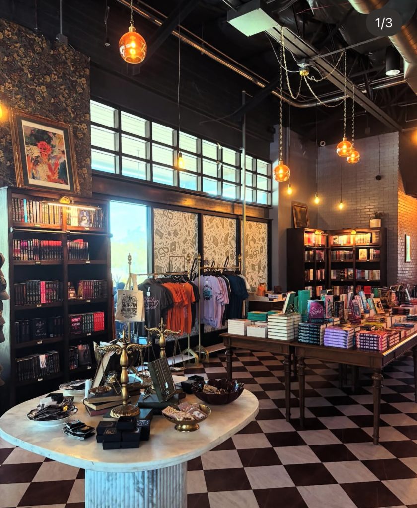 Inside a bookstore, yellow lights hang from the ceiling, and a large window sits at the back. Bookshelves are at the edges of the photo, and there are tables full of books and bookish items. A rack of shirts and tote bags sits underneath the window.