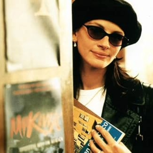 Julia Roberts from a movie still of Notting Hill wearing a black beret and sunglesses holding a book in a bookstore.